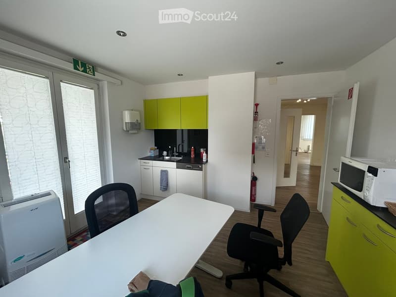 A furnished room with a table, chairs, a refrigerator, microwave, and a small kitchen area with a sink, faucet, and dishwasher. The room has wooden flooring and white walls.