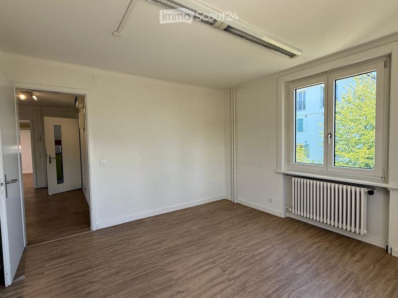 Empty room with wooden flooring, white walls, and a large window with a radiator below it. The room has a door leading to another area.