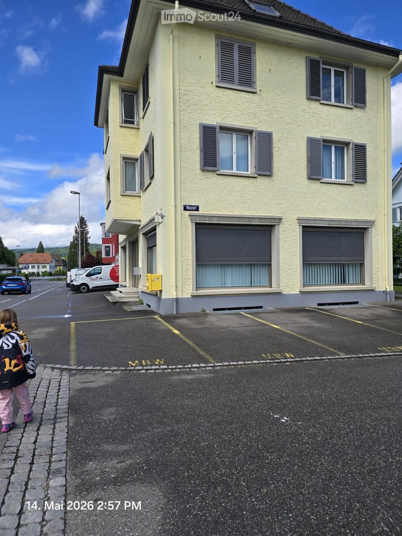 A multi-story residential building with light yellow walls, multiple windows with shutters, and a small entrance staircase. The building is situated next to a paved parking area with marked spaces.