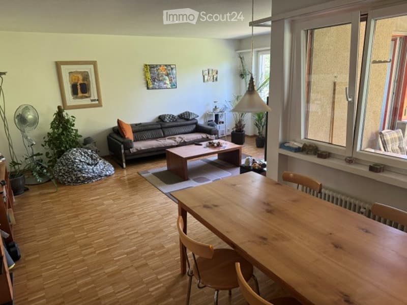 A modern living room with a grey sofa, wooden coffee table, dining table with chairs, potted plants, and a fan. The room features light-colored walls, wooden flooring, and large windows with blinds.