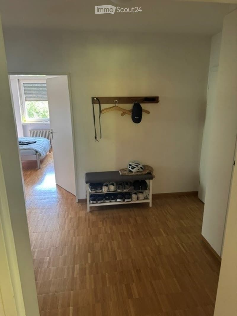 Empty hallway with wooden flooring, a shoe rack, and a coat hook. An open door leads to a bedroom with a bed visible.