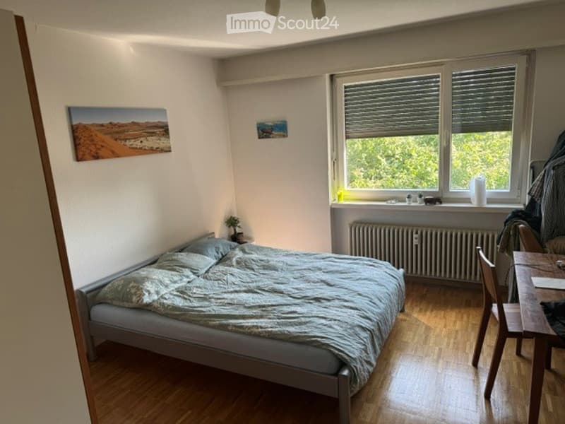 Cozy bedroom with a double bed, wooden floor, large window with blinds, small table and chair, radiator, and artwork on the walls.