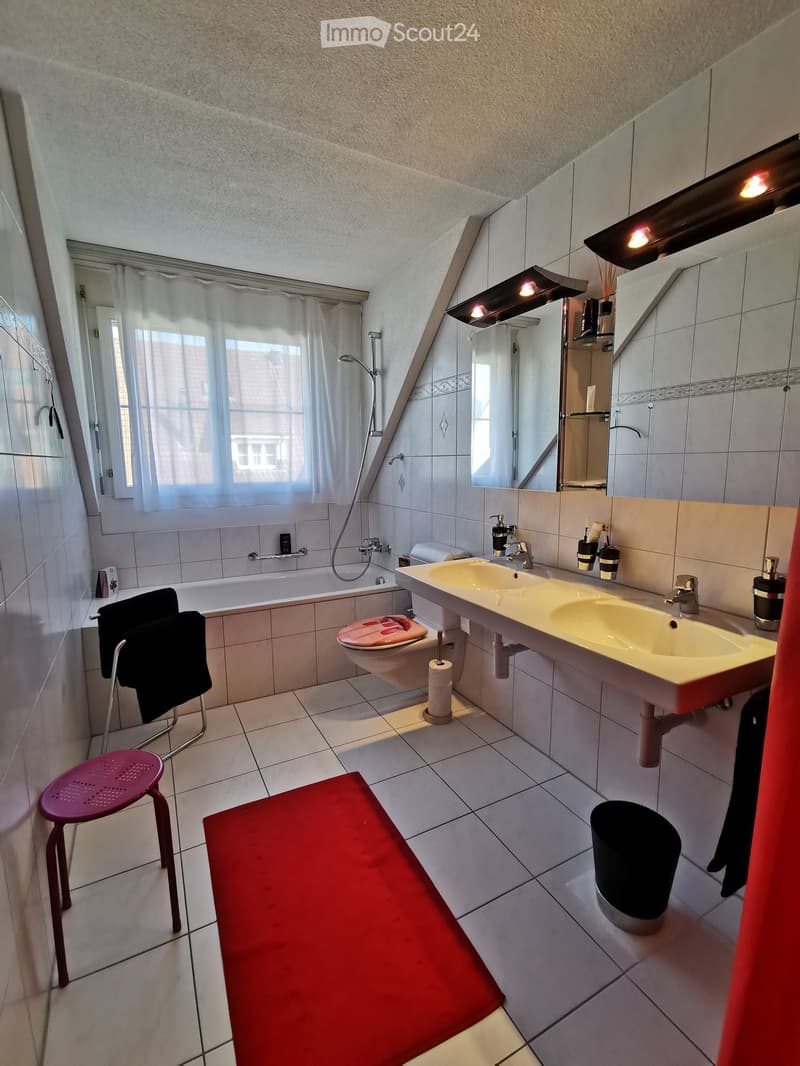 Modern bathroom with a double sink, bathtub, toilet, and a large mirror. Features include a red bench, a purple stool, and a window with white curtains.
