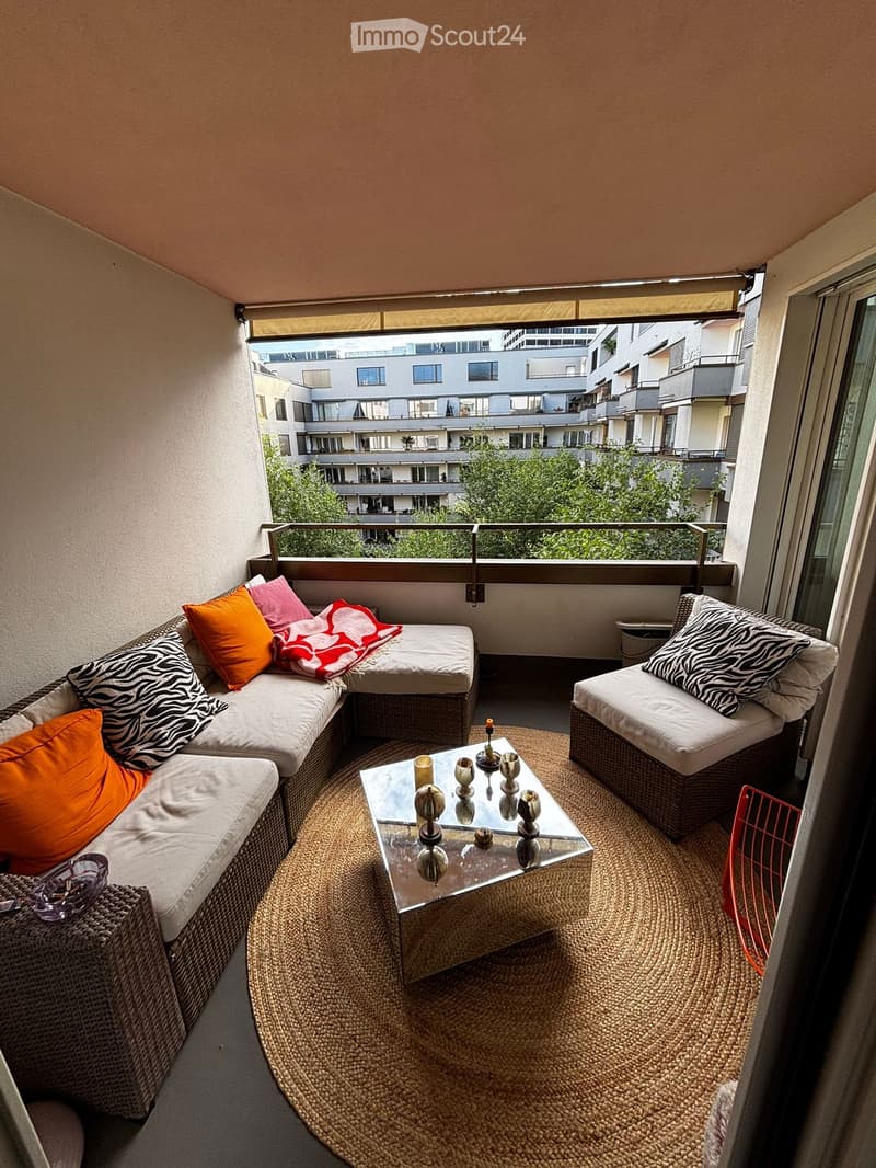 Cozy balcony with a corner sofa, coffee table, and chair, decorated with colorful cushions and a patterned throw, overlooking a cityscape with buildings and greenery.