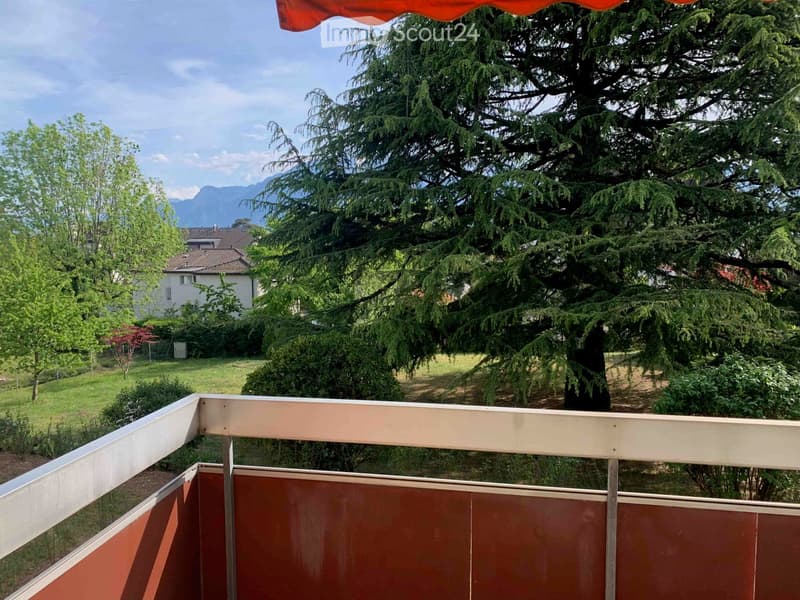 Balcony with wooden railing, overlooking a green garden and trees, with a distant view of mountains.