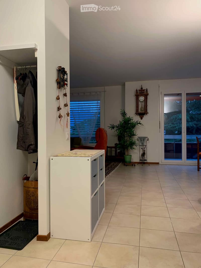 A well-lit hallway with tiled flooring, a white storage unit, a red chair, a potted plant, and a wall-mounted clock. A coat rack with hanging coats is visible on the left, and a sliding glass door leads to an outdoor area.