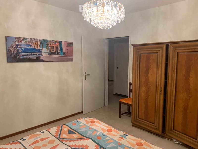 Bedroom with a bed, colorful bedspread, wooden wardrobe, chair, and a large picture on the wall.