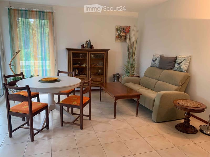 A well-furnished living and dining area with a beige sofa, wooden coffee table, round dining table with four chairs, a wooden cabinet, and decorative items including a plant and artwork.