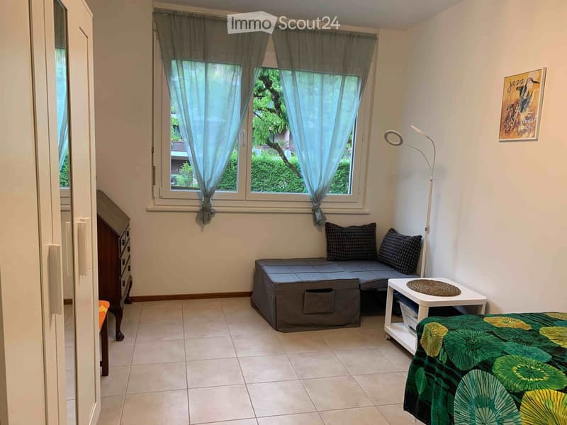 A small, cozy room with a bed, sofa, small table, and a floor lamp. The room has large windows with curtains and a view of greenery outside.