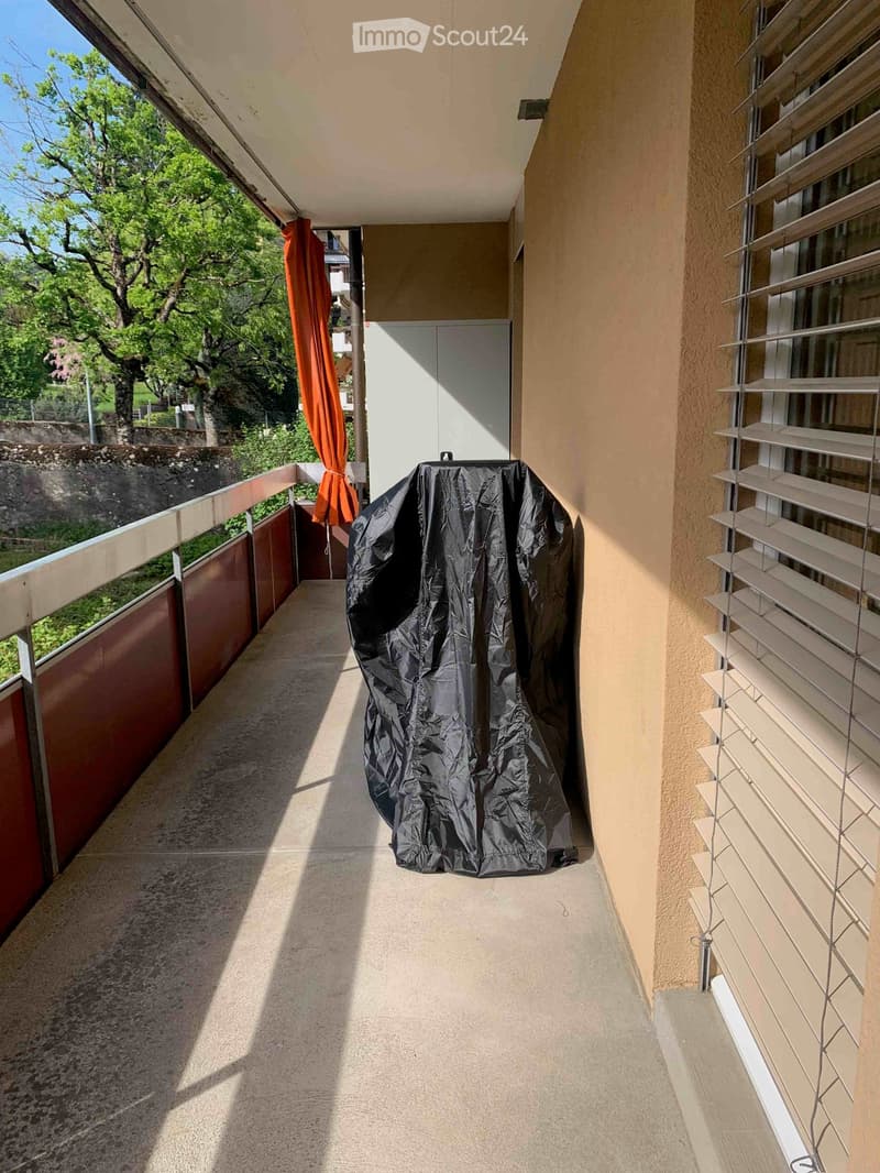 Balcony with beige walls, metal railings, and a covered outdoor item. The view shows greenery and a stone wall.
