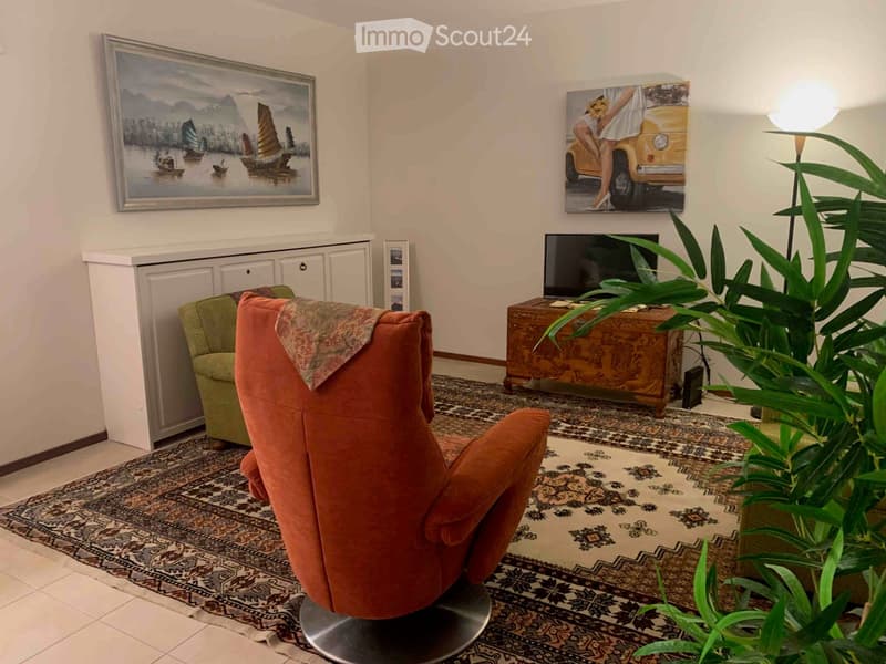 Living room with a patterned rug, orange sofa, green armchair, wooden cabinet, television, and decorative artwork on the walls.