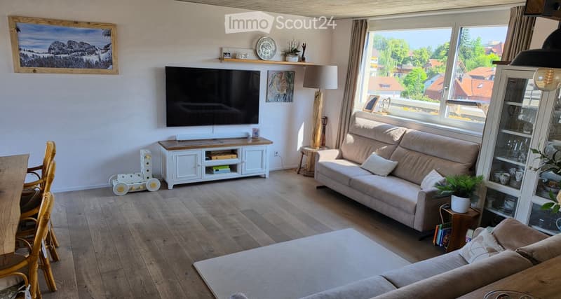 Bright living room with wooden flooring, large windows, a beige sofa, a wooden chair, a TV on a white cabinet, and decorative items including a lamp and a plant.