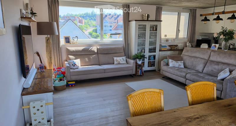 Bright living room with large windows, two sofas, a wooden dining table, and various decorative items including a guitar, a cat, and a vase with flowers.