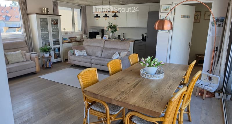 Bright living and dining area with wooden flooring, a large sofa, a dining table with chairs, a floor lamp, and a cabinet with glass doors.