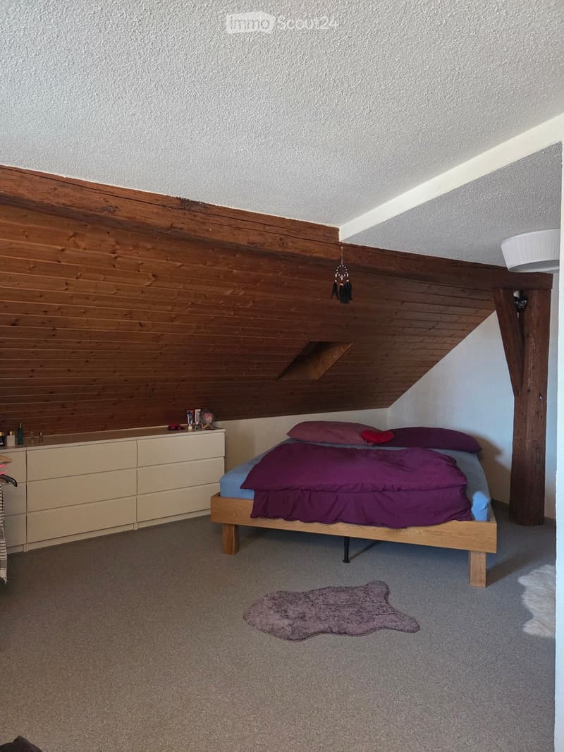Cozy bedroom with a wooden bed, purple bedding, a nightstand with a lamp, a drawer unit, and a small rug on the carpeted floor.