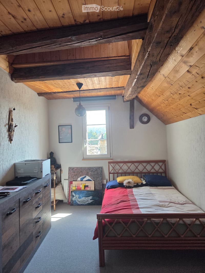 Cozy attic bedroom with wooden slanted ceilings, a bed with a colorful quilt, a nightstand with a lamp, a printer on a cabinet, and various artworks and decorations.