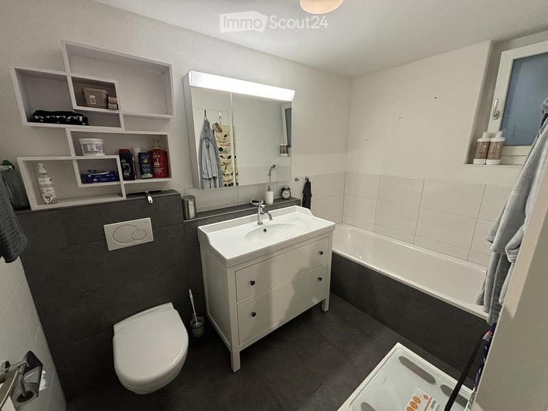 Modern bathroom with a white sink, mirror, toilet, bathtub, shelves, and a towel hanging on the wall.
