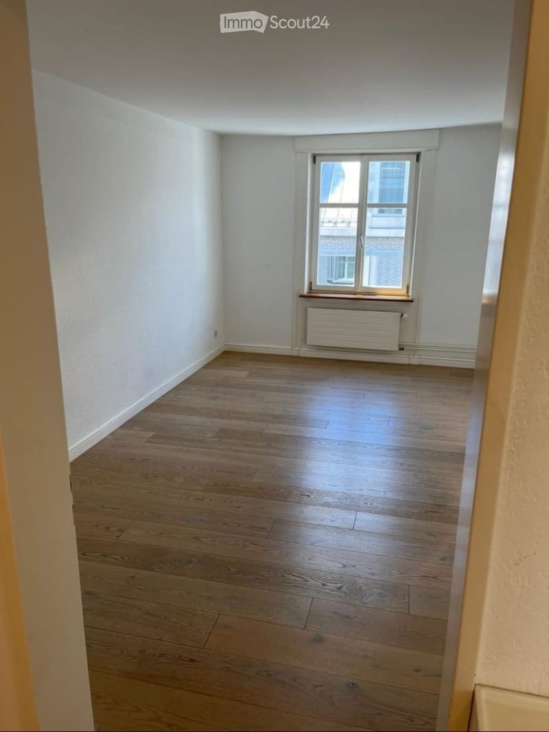 Empty room with wooden flooring, white walls, and a single window with a radiator below it.
