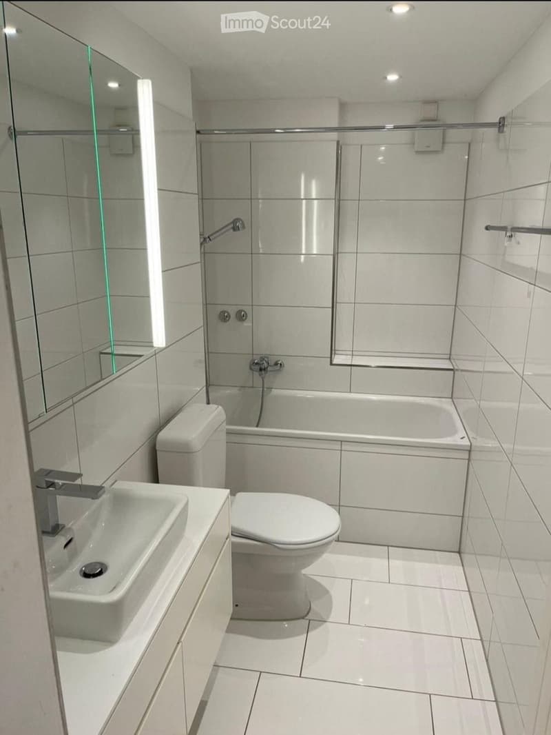Modern bathroom with white tiles, a bathtub, toilet, sink, and a large mirror above the sink. The shower area is enclosed with a glass door.