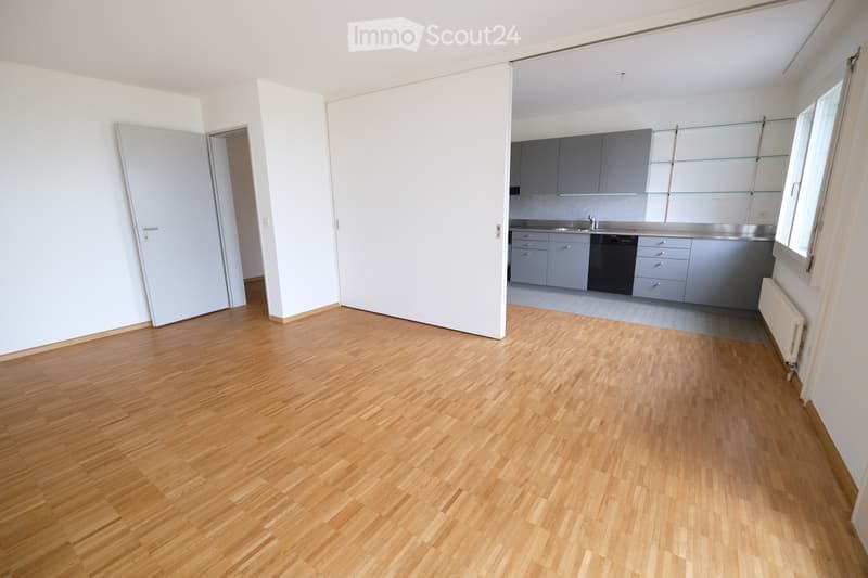 Empty room with wooden flooring, white walls, and an open doorway leading to a kitchen area with grey cabinets and stainless steel appliances.