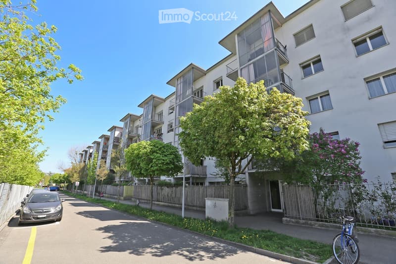Modern multi-story residential building with balconies and large windows, surrounded by trees and greenery. A parked car and bicycle are visible on the street.