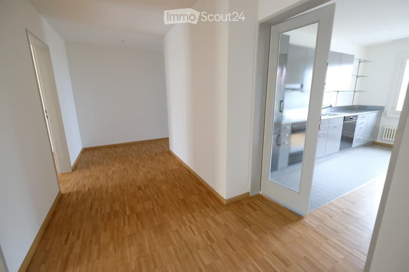 Empty hallway with wooden flooring and white walls, leading to a kitchen area with cabinets and a dishwasher.