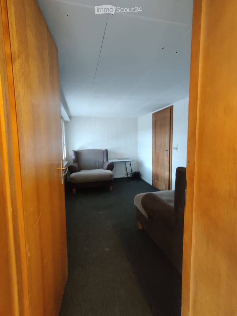 A small, furnished room with a sofa, a small table, and a partially visible bed, featuring wooden doors and a window.