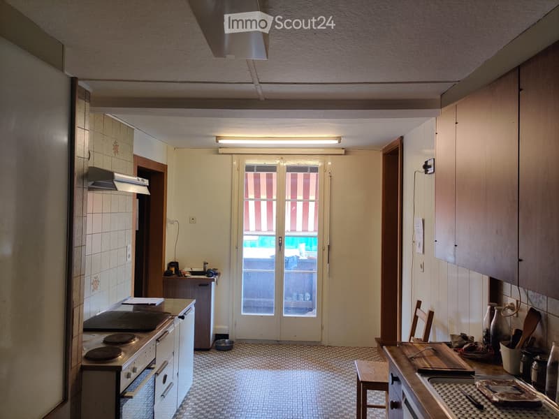 The kitchen features a stove with multiple burners, an oven, a refrigerator, a sink, a wooden table with a chair, and various kitchen utensils and items on the counter.