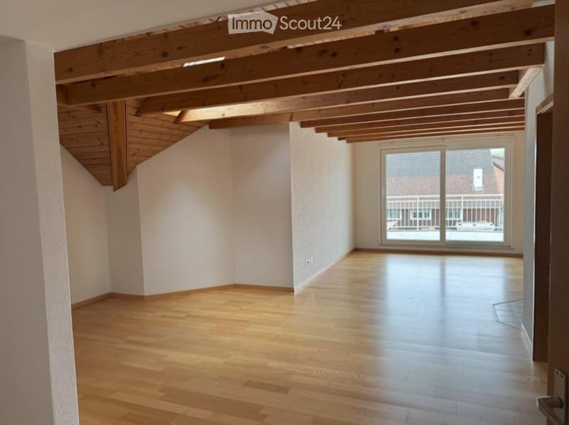Empty room with wooden flooring and exposed wooden beams on the ceiling. Large window with a view of the outside.