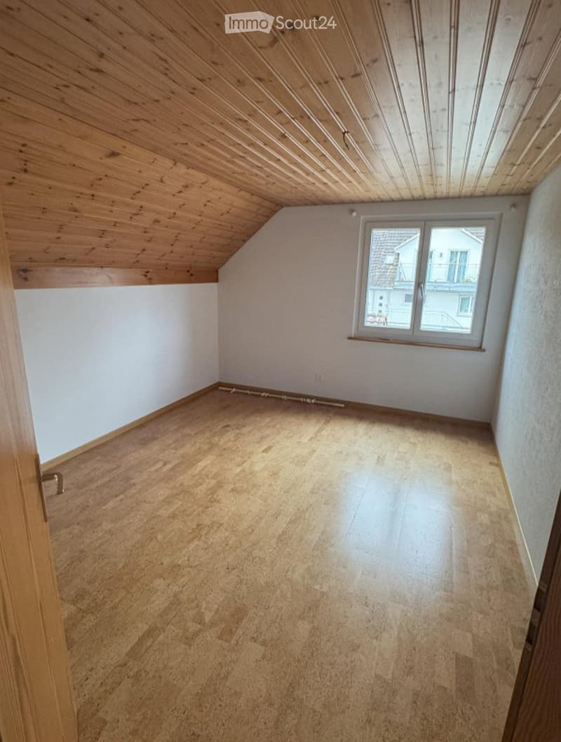 Empty room with wooden floor and slanted wooden ceiling, featuring a single window with white frame and a door with brass handle on the left side.