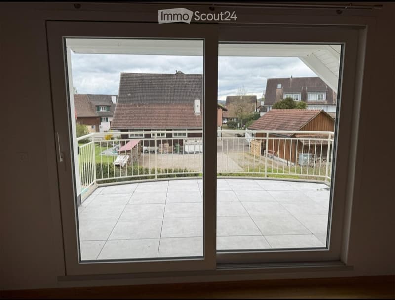 Balcony with tiled floor, white metal railing, overlooking a residential area with houses and greenery.