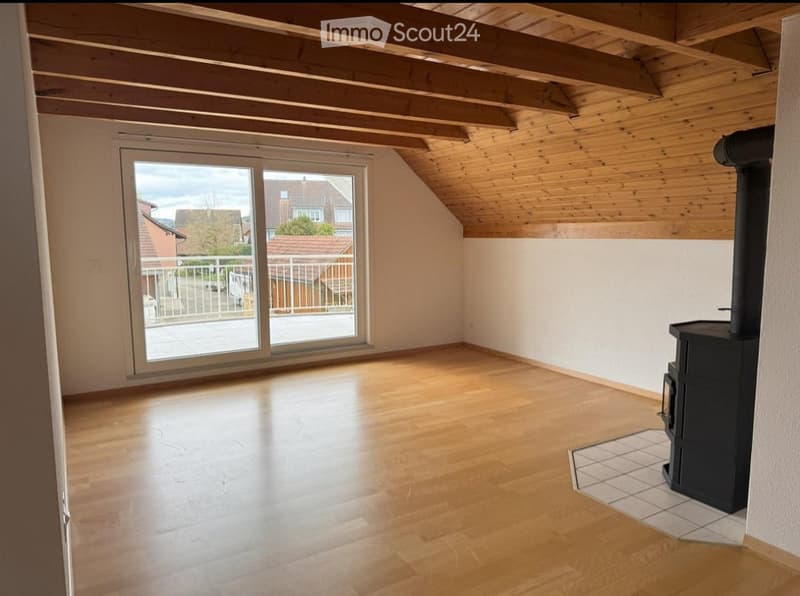 Empty attic room with wooden floors, exposed wooden beams, large window, and a fireplace.