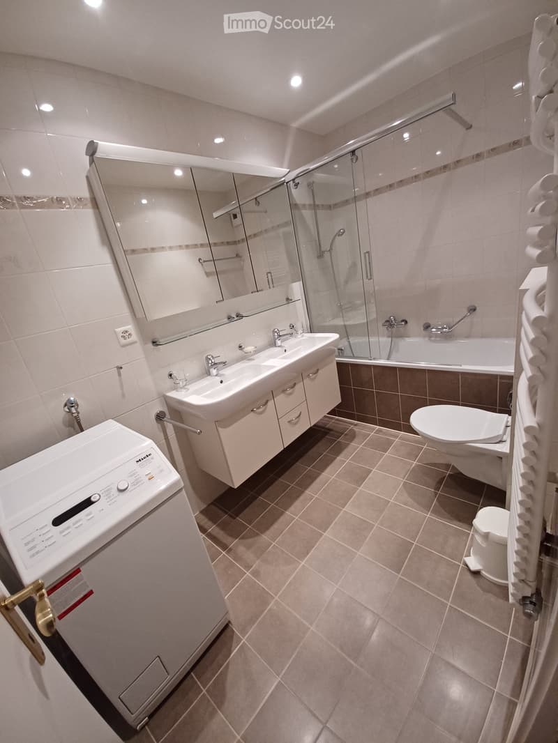 Modern bathroom with a double sink, mirror, bathtub, toilet, and washing machine. Features tiled walls and floor, and a glass-enclosed shower area.