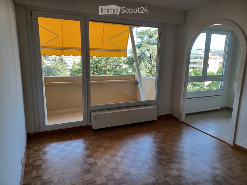 Empty room with wooden flooring, large window, and sliding glass door leading to a balcony with yellow awning.