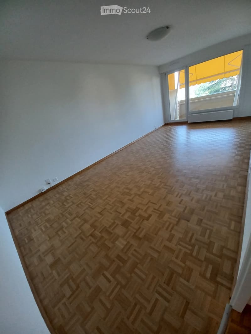Empty room with wooden herringbone flooring and white walls, featuring a large window with yellow blinds and a radiator below it.