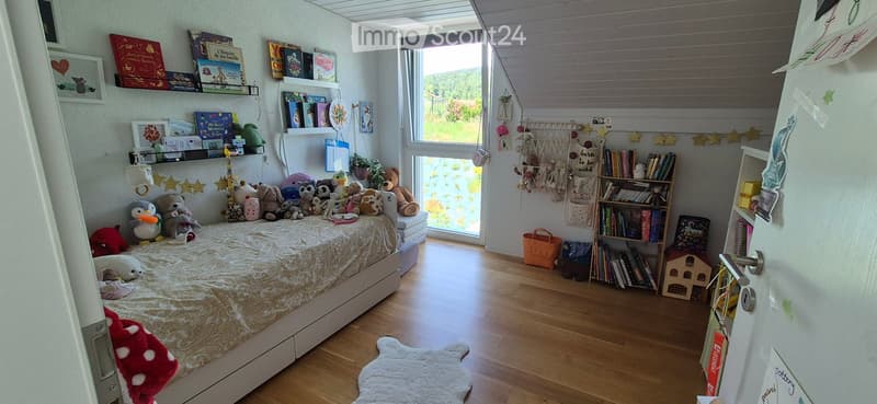 Cozy children's bedroom with a large bed covered in stuffed animals, bookshelves filled with books and toys, and a large window offering a view of greenery.