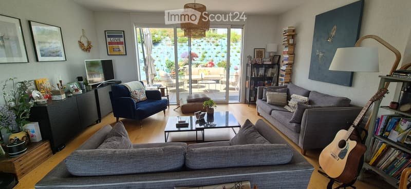Modern living room with large windows offering a view of a garden, featuring a grey sofa, blue armchair, coffee table, bookshelf, and a guitar leaning against the wall.