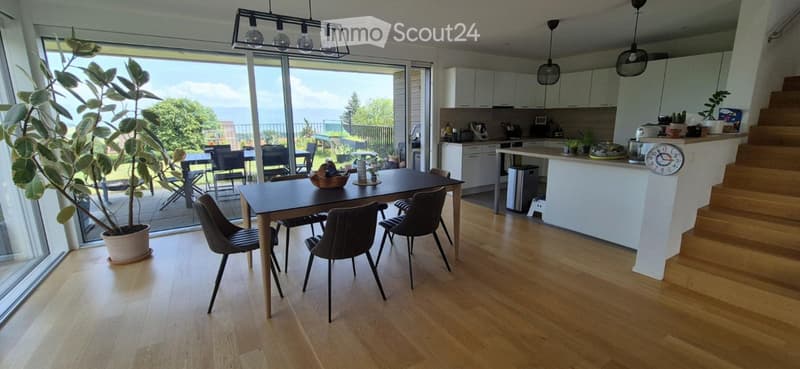 Bright, open-plan living and dining area with light wood flooring, large windows, a dining table with chairs, a kitchen island, and a potted plant near the window.