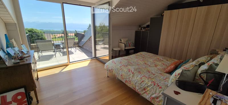 Bright bedroom with wooden flooring, large window offering a scenic view, bed with floral bedding, nightstand, chair, stool, and a small table with chairs on the balcony.