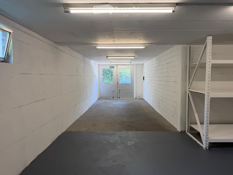 Empty storage area with white brick walls, concrete floor, fluorescent lighting, and a metal shelving unit on the right side.