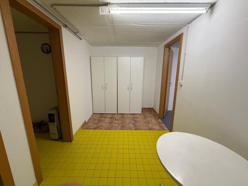 A small, narrow room with yellow tiled flooring, white walls, and a white table in the foreground. There is a white double-door cupboard and a small radiator visible.