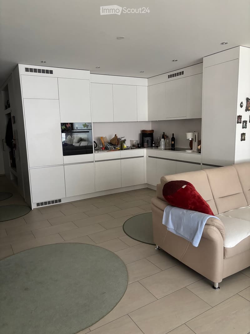 Modern open-plan living and kitchen area with white cabinets, a beige sofa with cushions, a round coffee table, and various kitchen appliances.