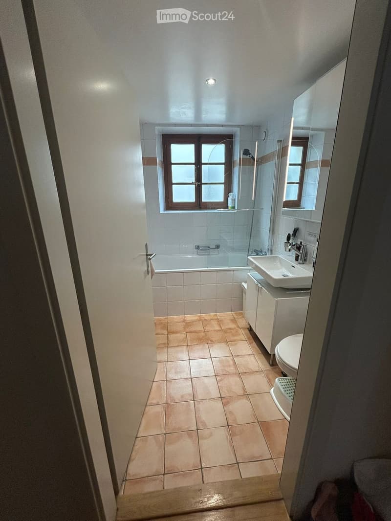Bathroom with tiled floor, bathtub, sink, toilet, and a glass-enclosed shower area. The room has a window with wooden frames and a small cabinet next to the sink.