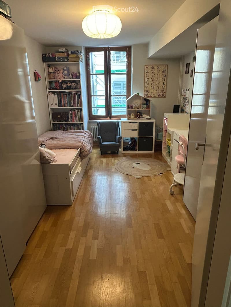 Cozy children's bedroom with wooden flooring, a single bed with a pink blanket, a small armchair, a bookshelf filled with books and toys, a storage unit, a round rug, a small desk with a chair, and a toy house on a shelf.