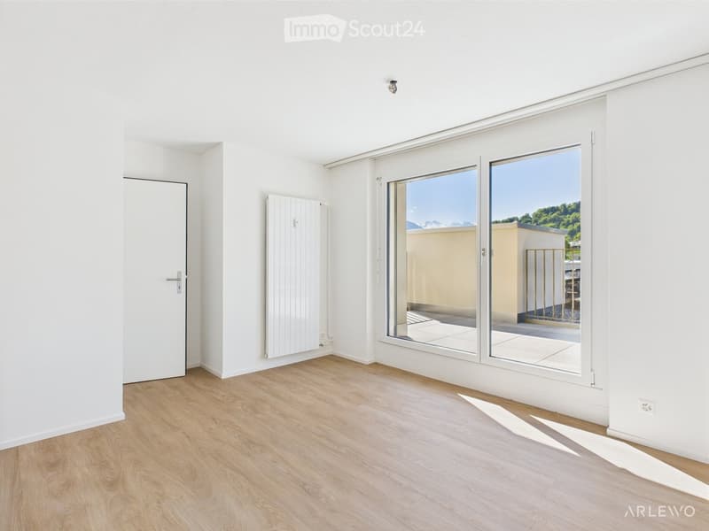 Empty room with light wooden flooring, white walls, a white door, a radiator, and large sliding glass doors leading to a balcony with a view of greenery and mountains.