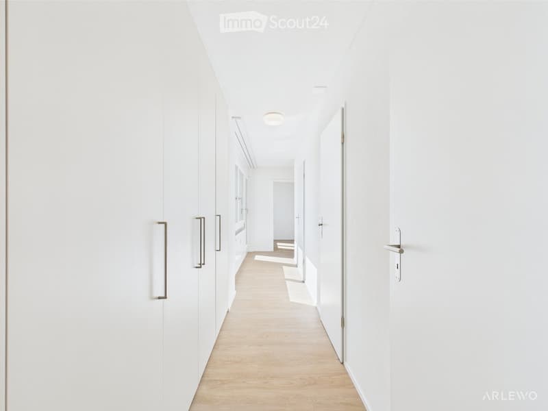 A clean, modern hallway with white walls, light wooden flooring, and recessed ceiling lighting. The hallway features several closed doors with sleek metallic handles, leading to other rooms.