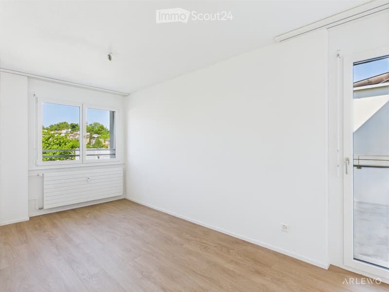 Empty room with white walls, wooden flooring, large windows, and a sliding glass door leading to a balcony.