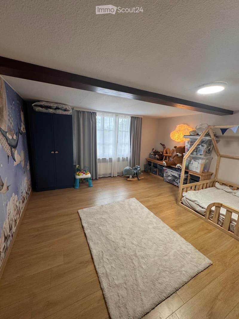 Child's bedroom with wooden floor, large window with curtains, bed, wardrobe, toys, and a shelf with storage boxes.