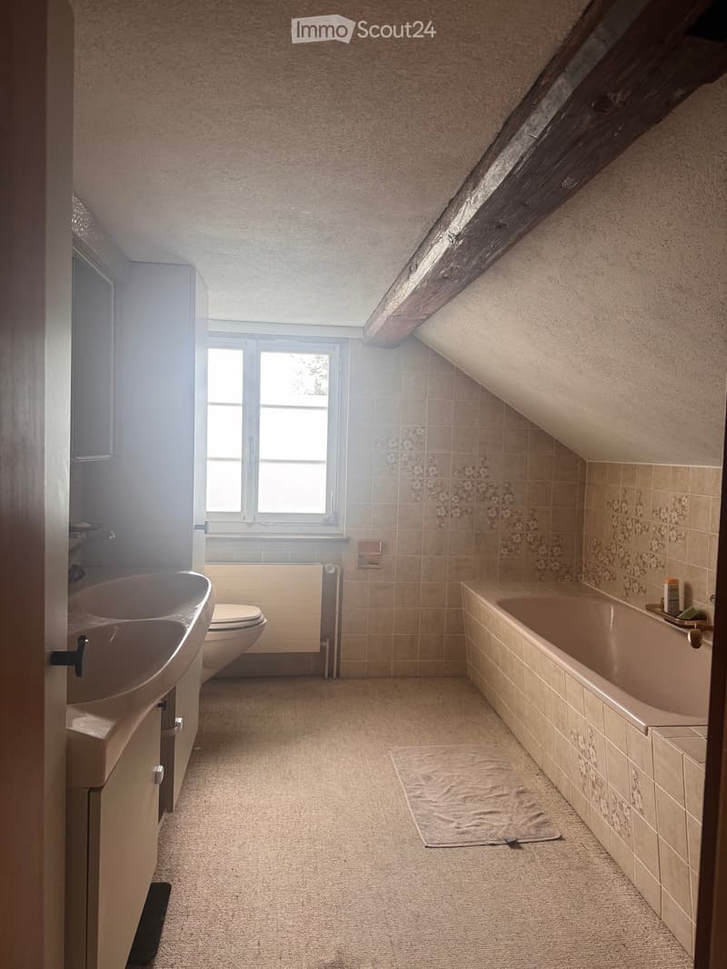 Bathroom with a sink, toilet, bathtub, and a towel on the floor. The room has beige tiles and a slanted wooden ceiling.