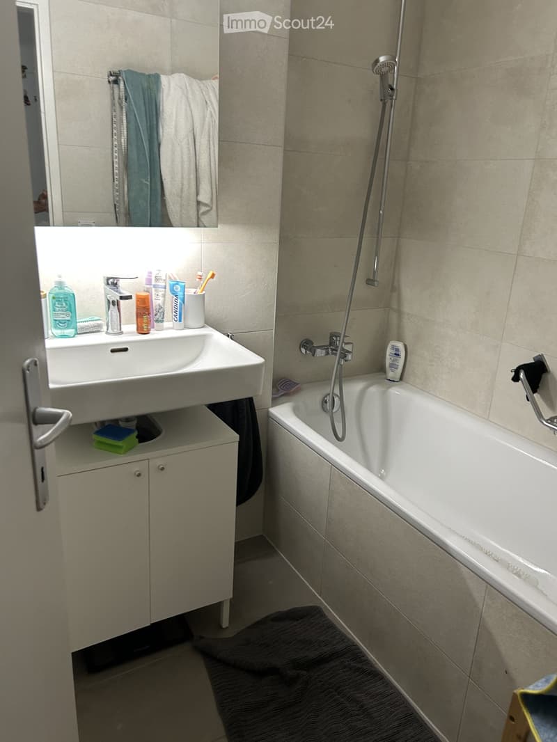 Modern bathroom with a white sink, bathtub, mirror, and various toiletries on the sink counter. Beige tiles and a dark bath mat are visible.